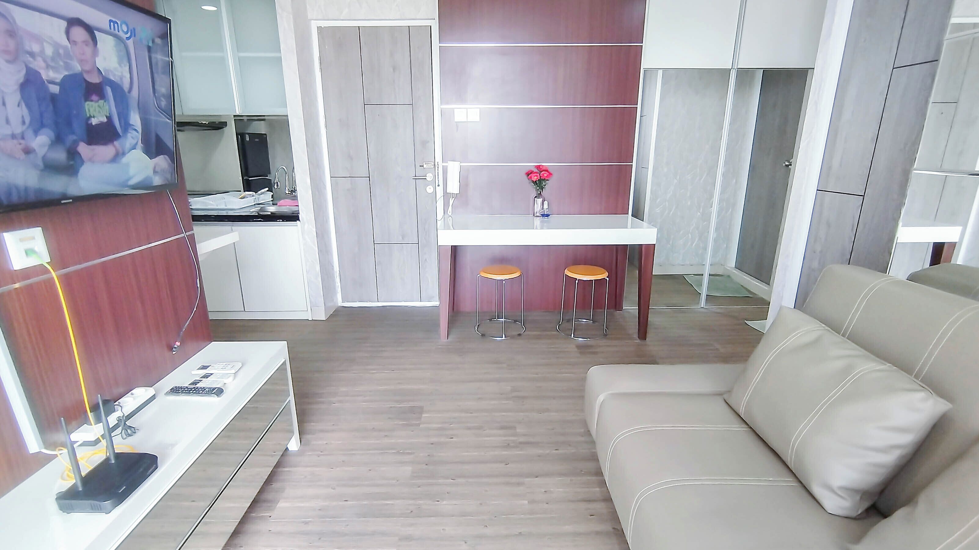 A modern living room with a beige sofa, a small dining area with stools, and a TV mounted on the wall.