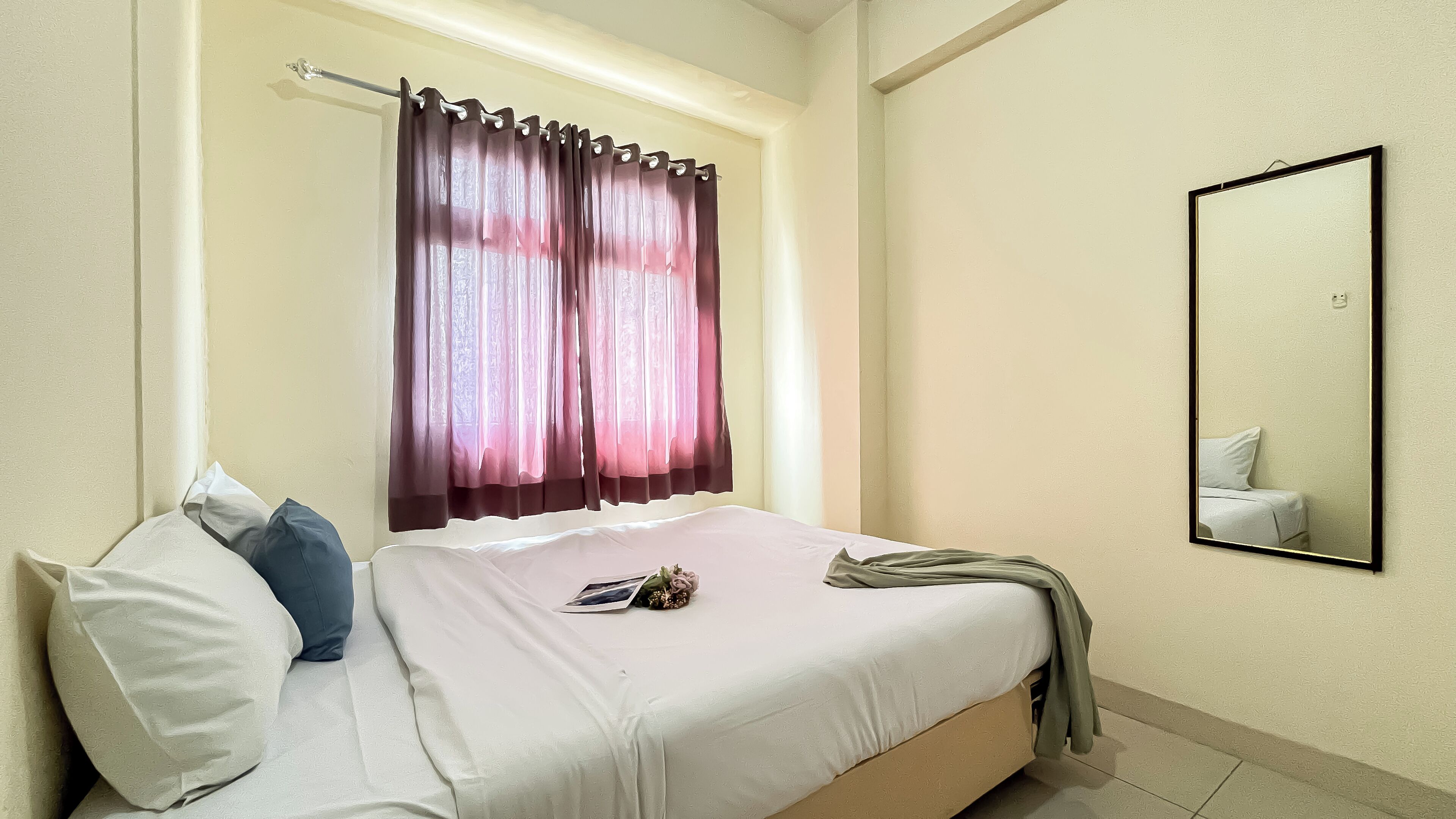 Un dormitorio con una cama, almohadas, una ventana con cortinas, un espejo y una mesita con un libro y flores.