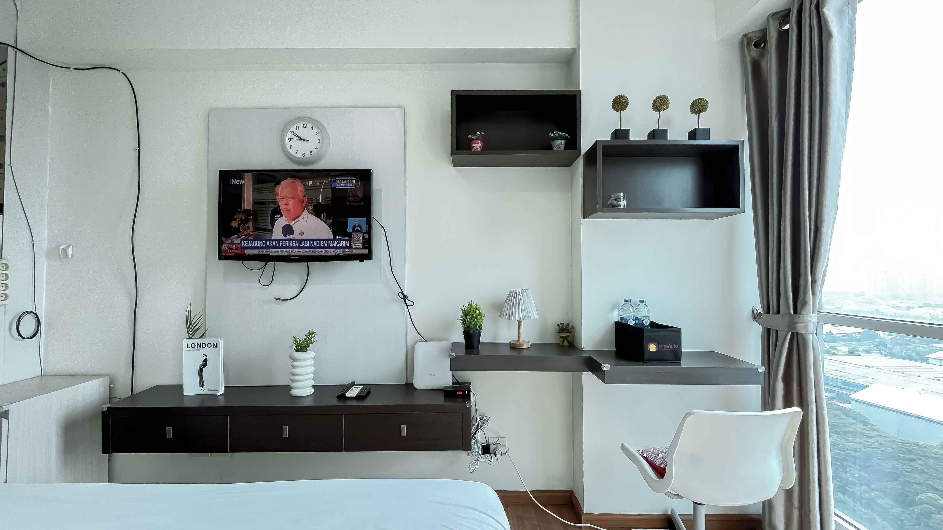 A hotel room with a bed, a TV mounted on the wall, a desk with a lamp, a small shelf with decorative items, and a view of the city through the window.