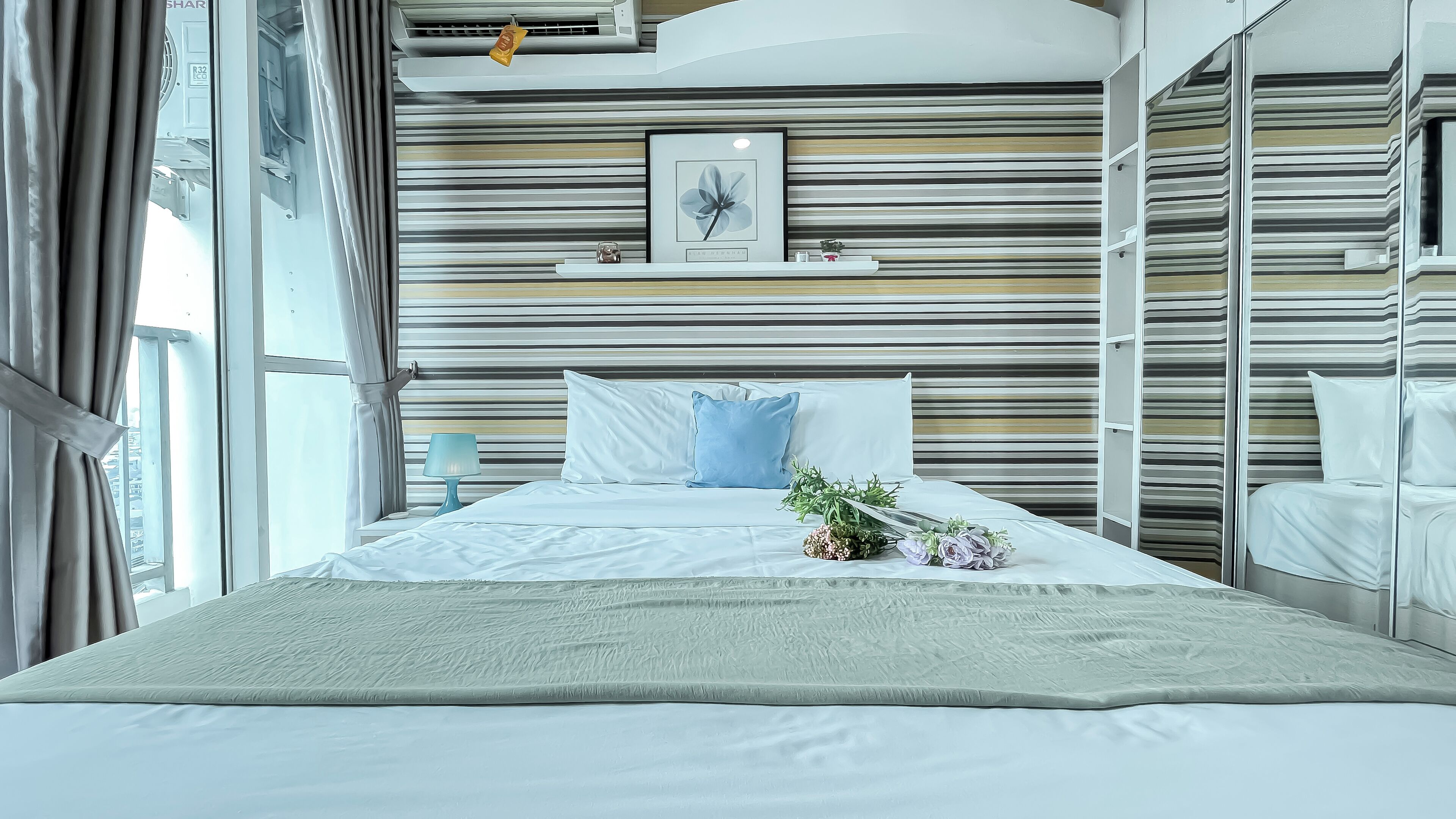 Une chambre moderne avec un grand lit, une table de chevet avec une lampe, un tableau encadré au mur et une fenêtre avec des rideaux.