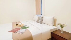 A neatly made bed with white linens, a bedside table with a vase of flowers, and a magazine on the bed. - Warm And Cozy 1Br With Makeup Room At Meikarta Apartment (Cikarang)