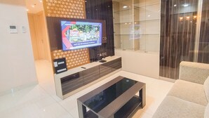 A modern living room with a flat-screen TV mounted on a wall, a glass cabinet, a sofa, and a coffee table. - Warm 3Br Direct Access To Mall La Riz Supermall Mansion Apartment (Surabaya)