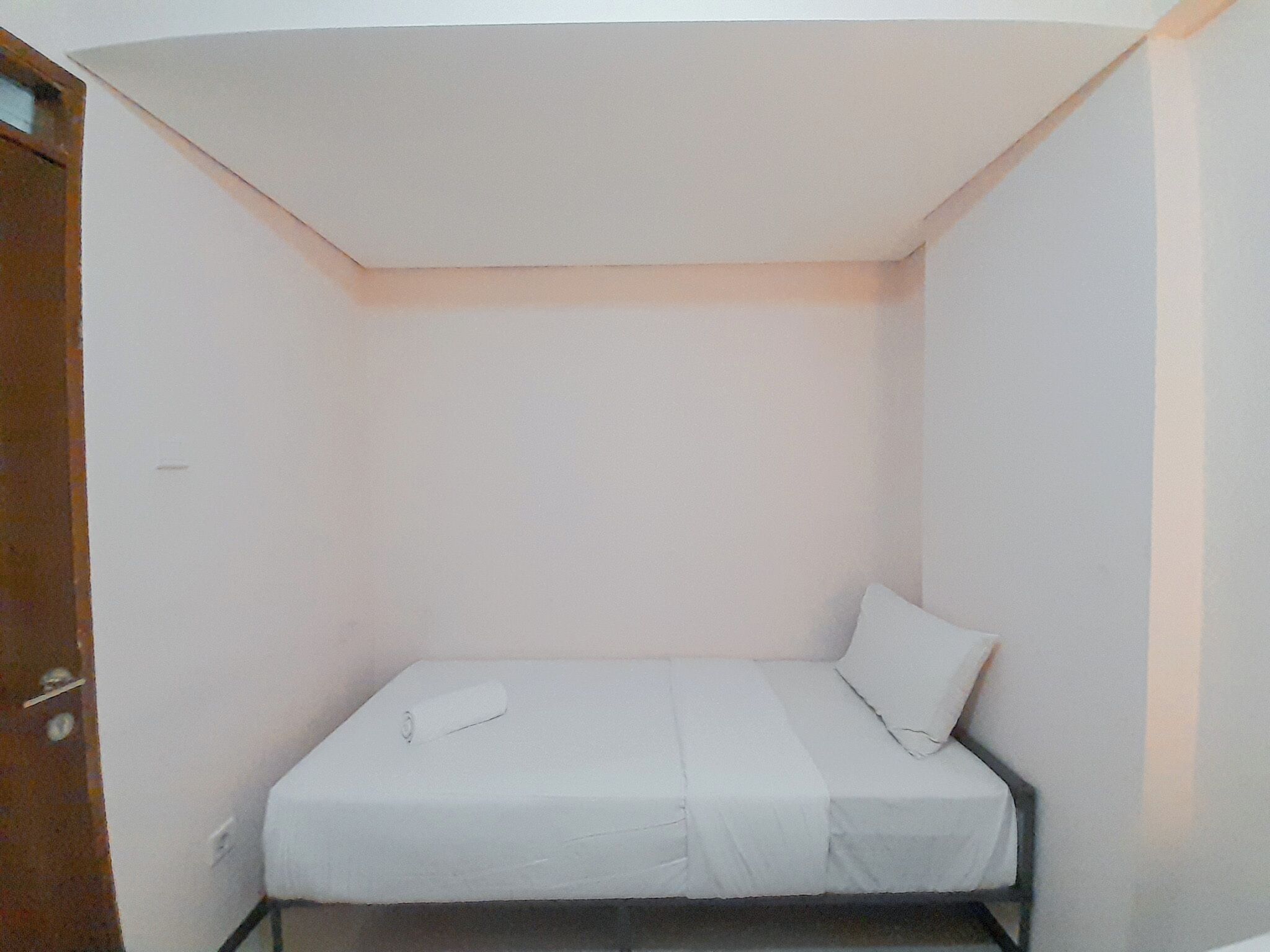 A small, single-bed room with a white mattress and pillow, a wooden door, and a white ceiling.