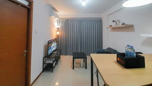 Un espace de vie compact avec une porte en bois, un téléviseur posé sur un meuble, une table à manger avec une bouteille d’eau, une petite table noire et un rideau gris.