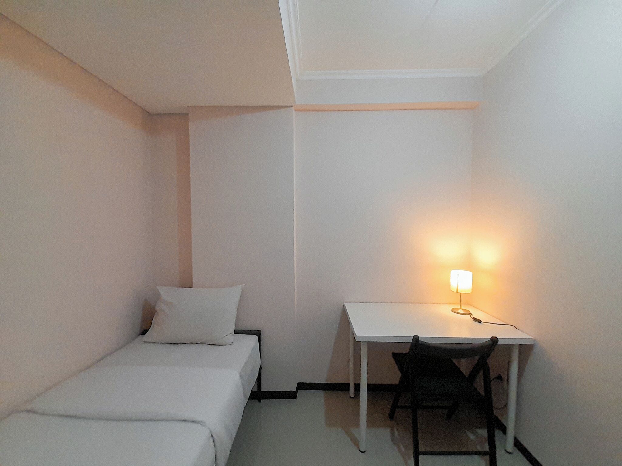Une petite pièce blanche, avec un lit simple, un bureau et une chaise.