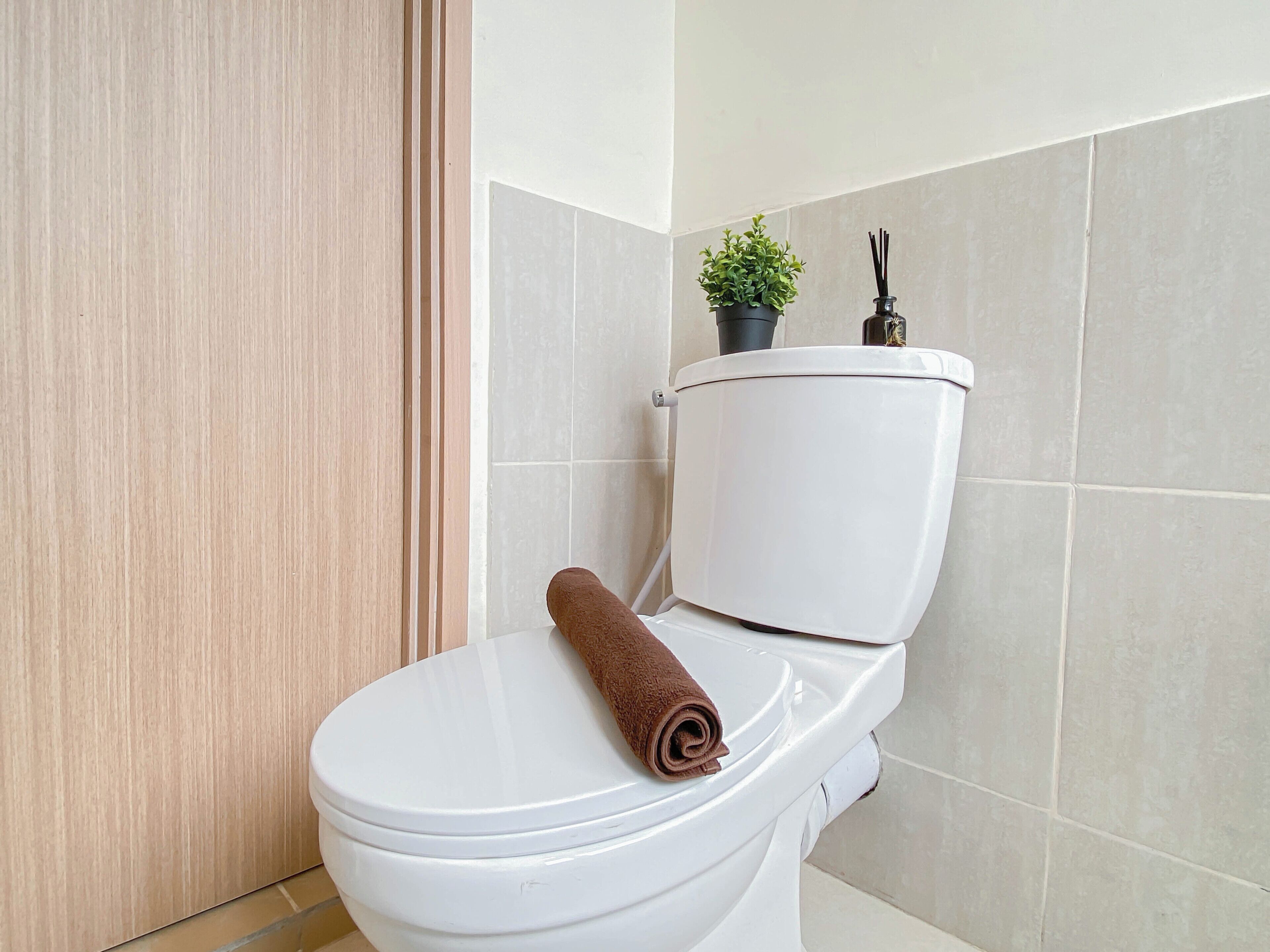 A white toilet with a brown towel on the seat, a small potted plant, and a reed diffuser on the tank.