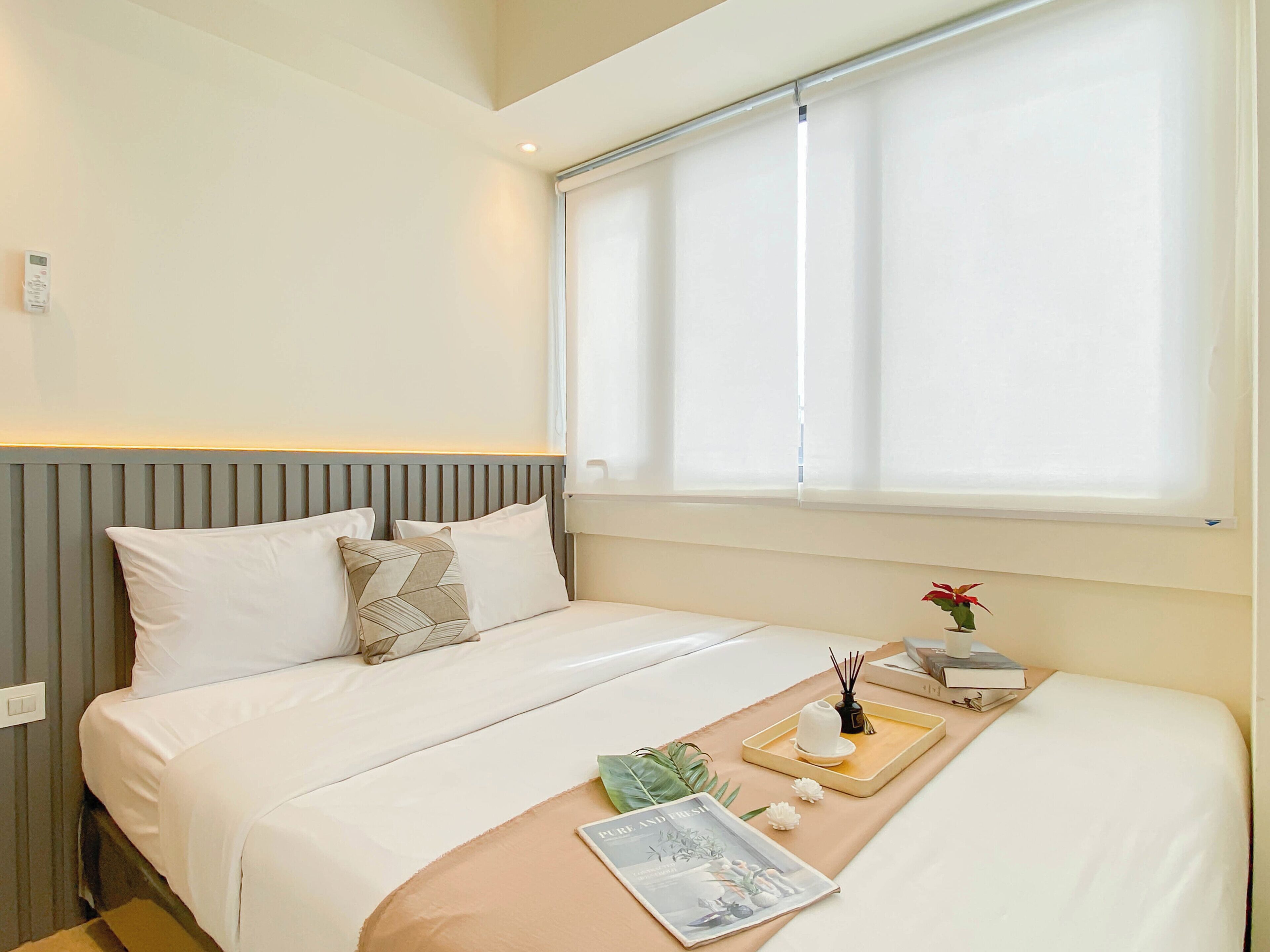 Een netjes opgemaakt bed met witte lakens en een beige plaid, een nachtkastje met een dienblad waarop een boek ligt, een vaas met een rode bloem en een kleine plant.