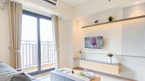 A modern living room with a sofa, a coffee table, and a TV mounted on the wall. There are two shelves with decorative items and a window with curtains. - Modern Style 2Br Apartment At Meikarta (Cikarang)