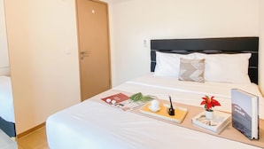 A hotel room with a bed, a bedside table with a book, a vase with a flower, and a door. - Simple Look 2Br Apartment Meikarta (Cikarang)