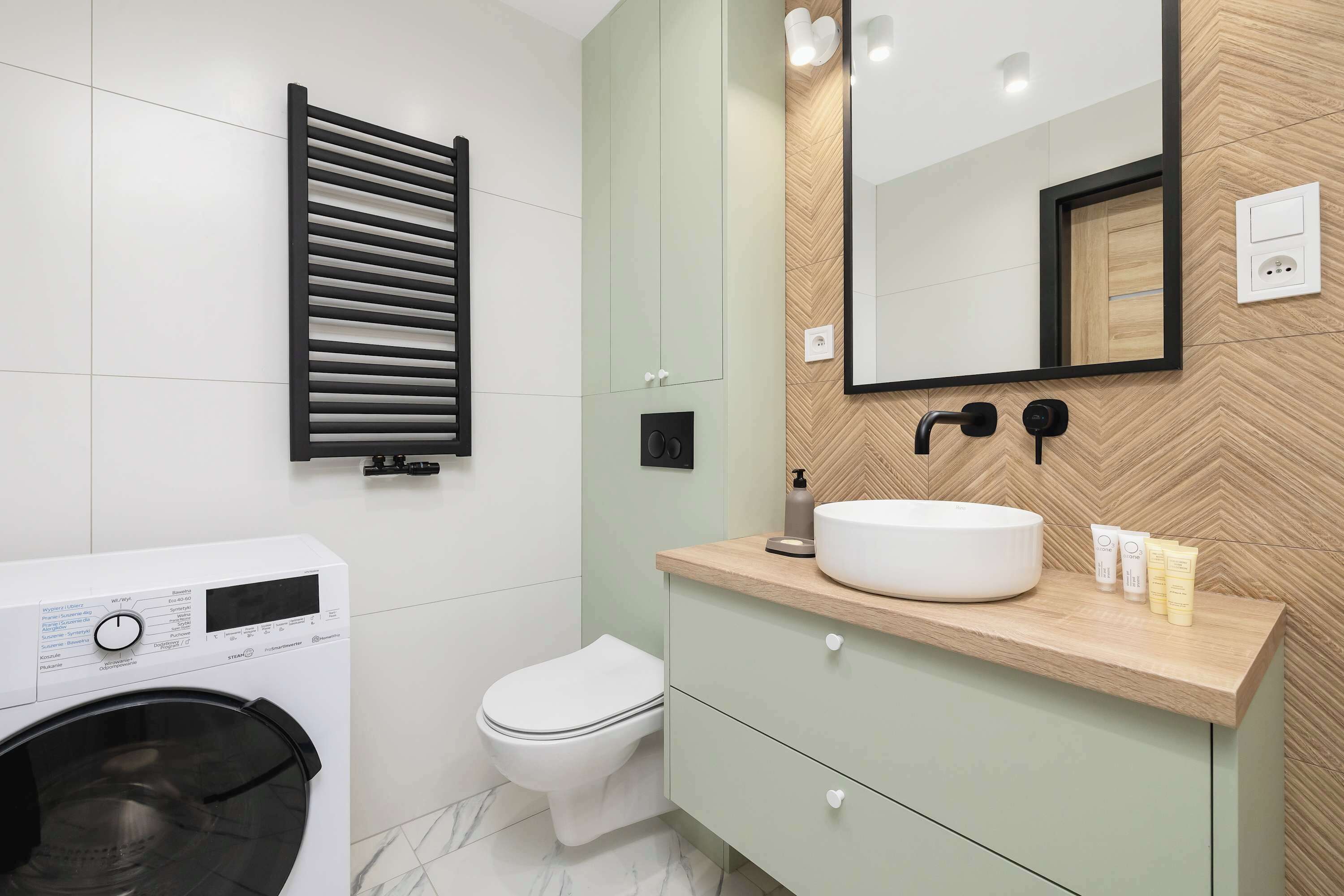 A modern bathroom with a built-in washing machine, a toilet, a vanity with a mirror, and a herringbone-patterned wall.