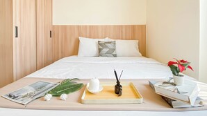 Una cama bien hecha con ropa de cama blanca, una bandeja con una botella pequeña y un objeto decorativo, una revista y una planta en maceta al lado.