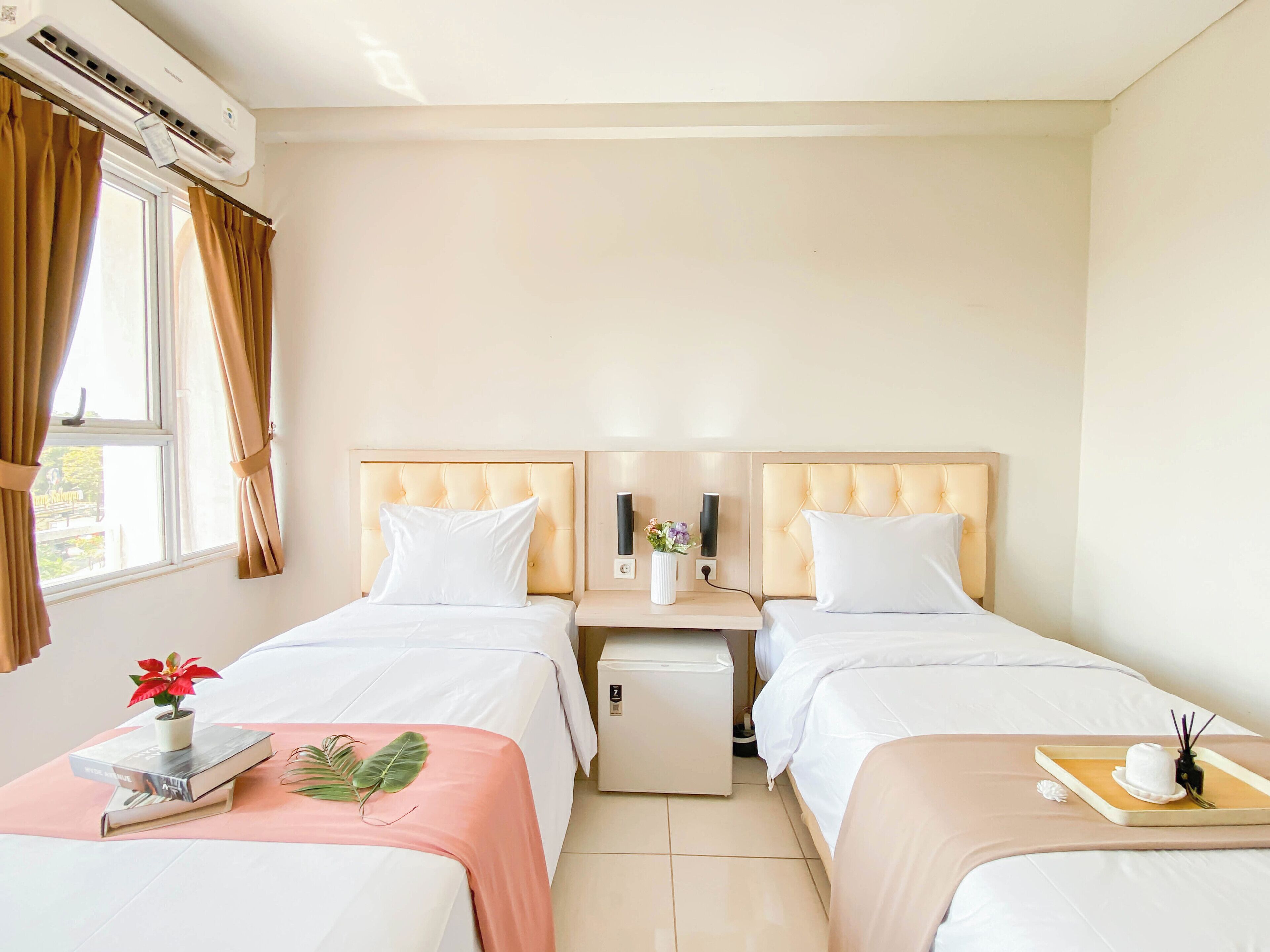 Camera d'albergo con due letti, un tavolino con una pianta e dei libri, un comodino con una lampada e un vaso, e una finestra con tende.