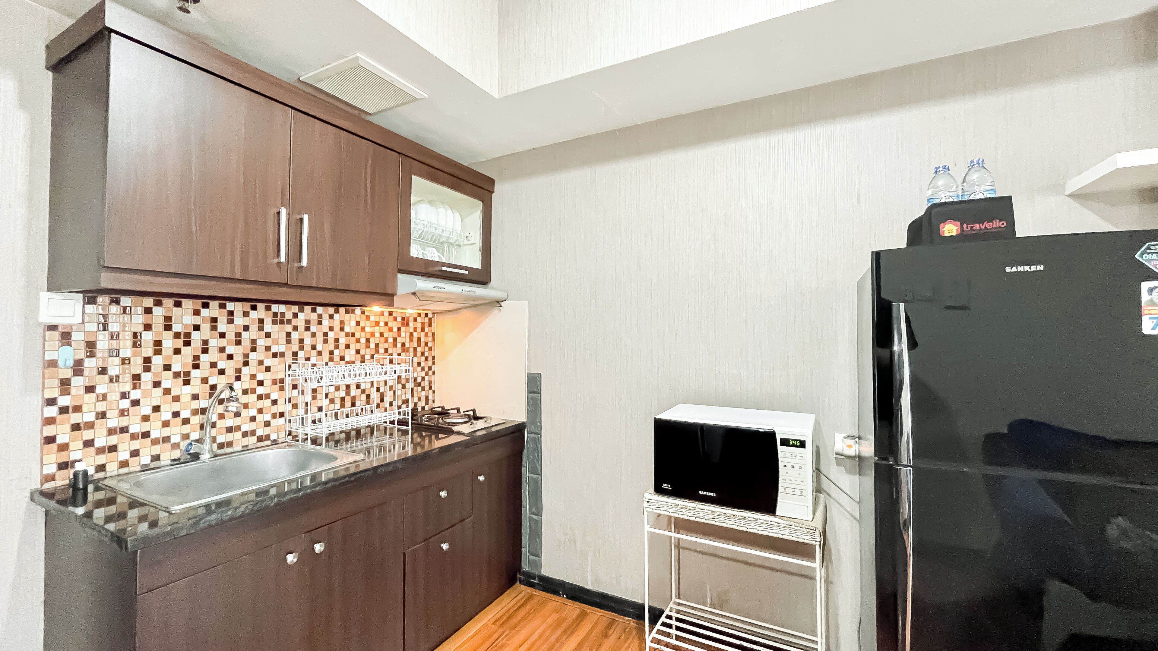 A compact kitchen with dark wooden cabinets, a tiled backsplash, a stainless steel sink, a microwave, and a black refrigerator.