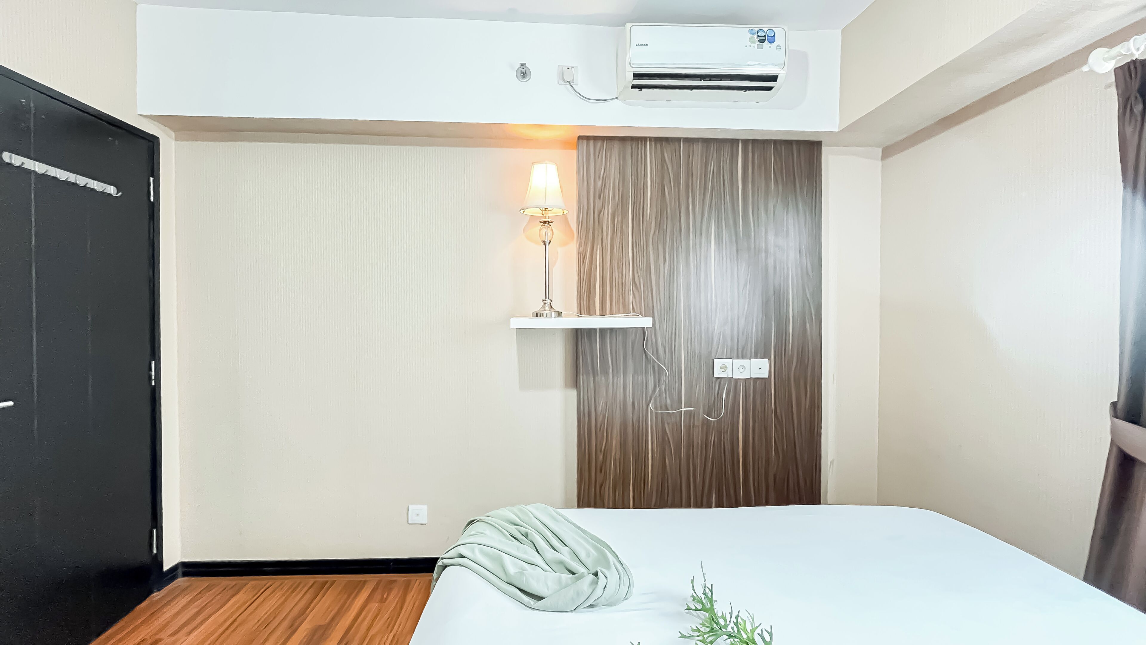 A hotel room with a bed, a wall-mounted air conditioner, a bedside lamp, and a wooden panel wall.