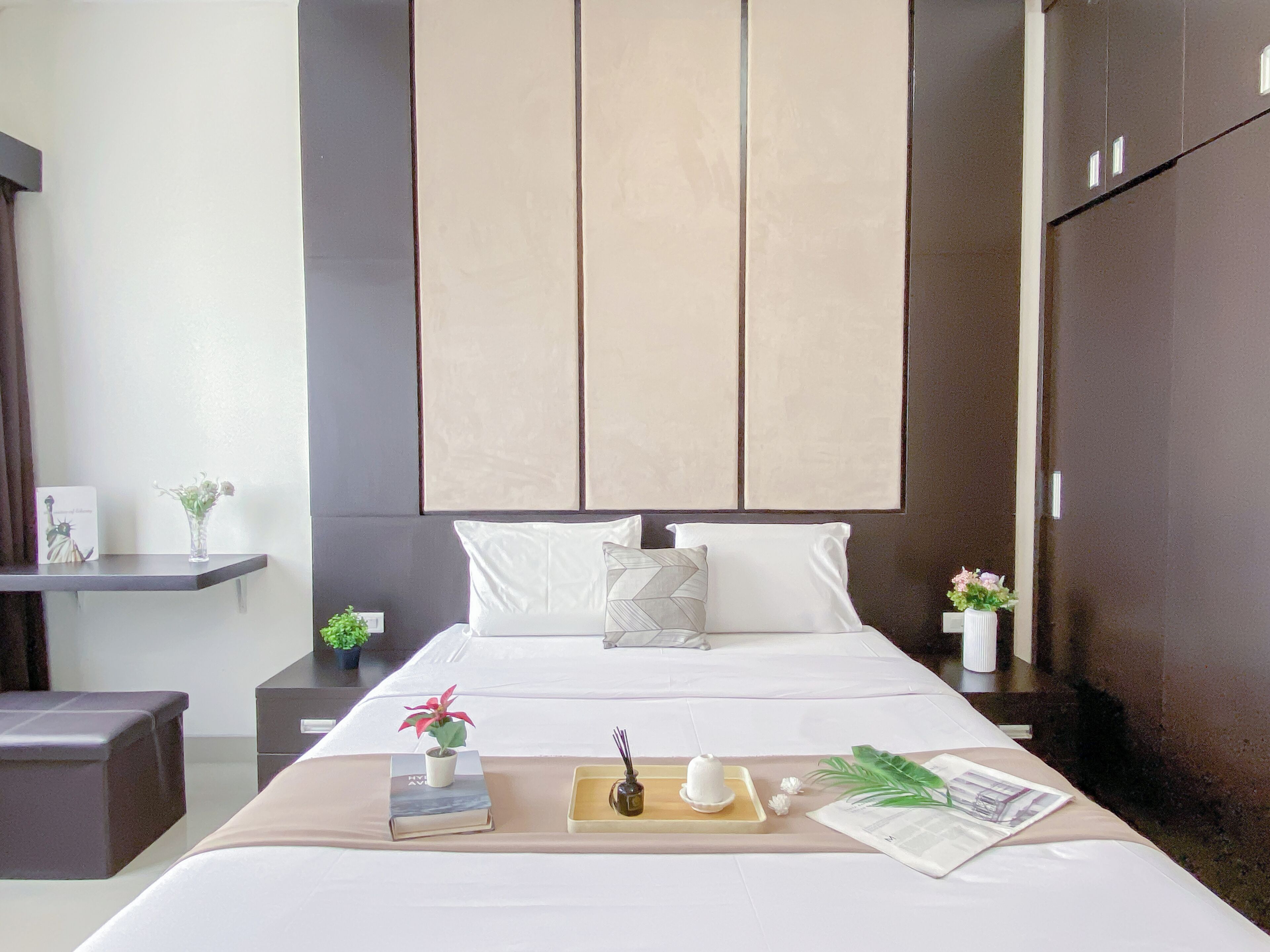 A modern hotel room with a large bed, a bedside table with a plant, a small table with a book and a candle, and a large wardrobe.