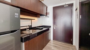 A compact kitchen with wooden cabinets, a stainless steel refrigerator, a black countertop, and a sink. - Comfy Rustic Studio Apartment At Casa De Parco (Pagedangan)