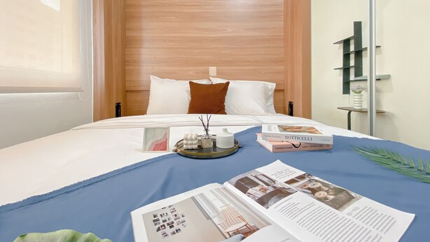 A hotel room with a wooden headboard, a bed with white and brown pillows, a blue bedspread, and a tray with books and a small vase. - Compact 2Br Apartment Direct Access To Mall At Green Pramuka City (Jakarta)