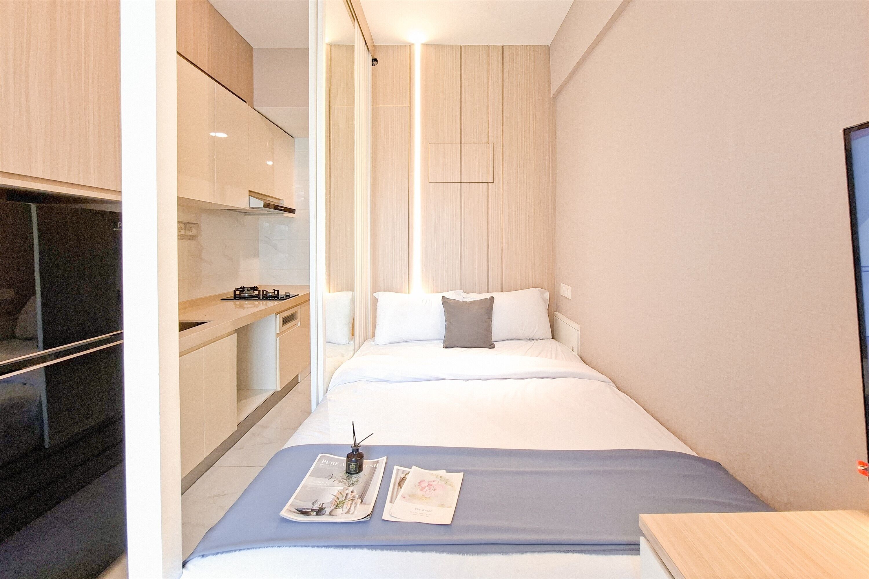 Habitación de hotel compacta con kitchenette, una cama con cubrecama azul y una mesita de noche pequeña con un libro y un control remoto.