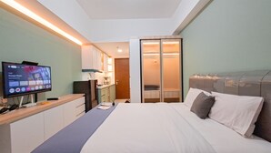 A modern hotel room with a bed, a desk with a computer, a TV, and a sliding door closet. - Simple Neutral Toned Studio Room At Parkland Avenue Apartment (South Tangerang)