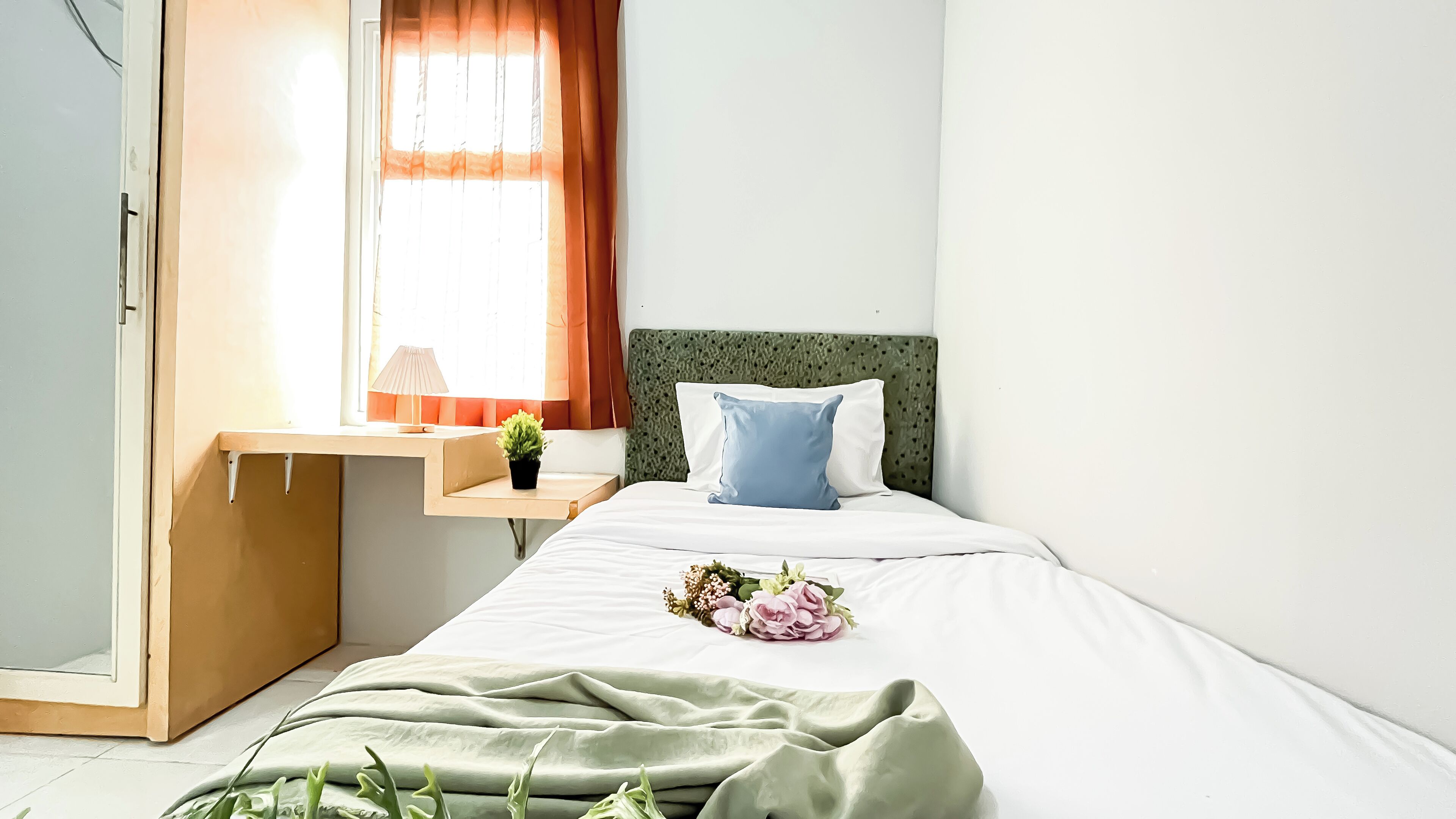 Un dormitorio ordenado con una cama, una mesita de noche con lámpara, una planta pequeña y una ventana con cortinas.