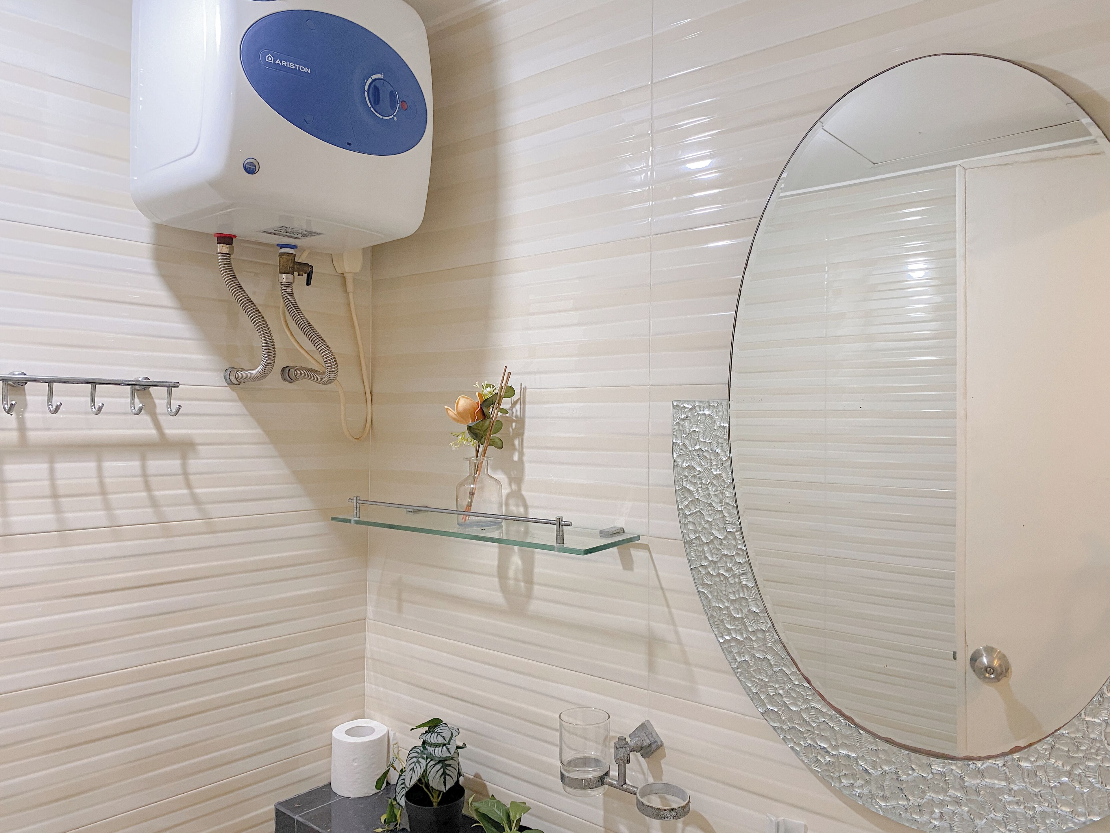 Ein Badezimmer mit Toilettenpapierhalter, einer Glasablage mit einer Vase, einem runden Spiegel mit dekorativem Rahmen und einer Duschbrause.
