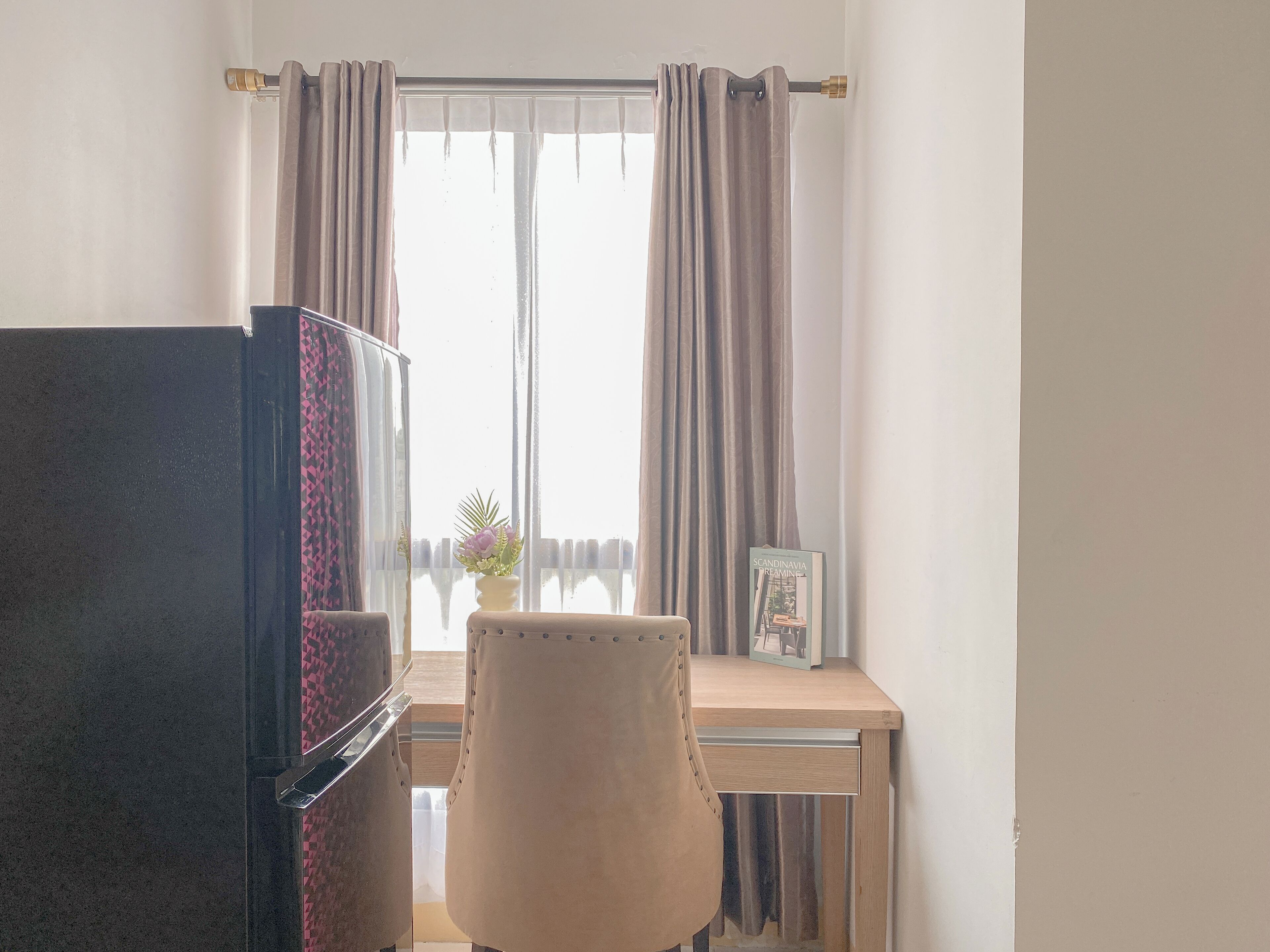A room with a refrigerator, a chair, a table with a picture frame, and curtains.