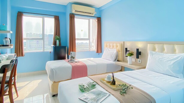 Ein Hotelzimmer mit zwei Betten, einem Schreibtisch und einem Sessel. Es gibt Fenster mit Vorhängen, eine Klimaanlage und einen Blick auf Gebäude.