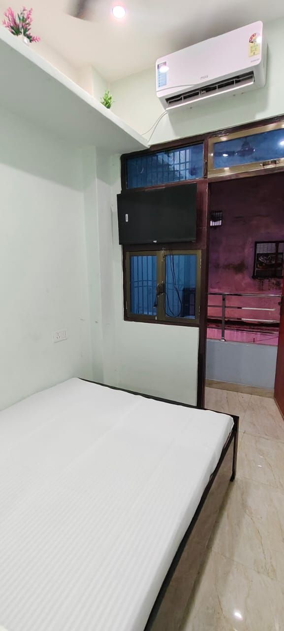 Een kamer met een enkel bed, een airconditioner, een raam met een blauw gordijn en een kleine plant aan de muur.