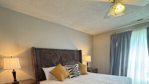A neatly made bed with a headboard, two bedside lamps, a ceiling fan, and a window with curtains. - Cozy 5BR / 2.5BA- Brick Home in Olive Branch (Olive Branch)