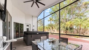 A modern outdoor dining area with a glass table and chairs, a hot tub, and a view of palm trees. - Modern Naples Home | Golf Course • Heated Pool & Spa (Naples)