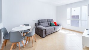 Un salón moderno con un sofá gris, una mesa de comedor redonda con sillas y un amplio ventanal que proporciona luz natural.
