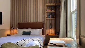 A neatly arranged hotel room with a bed, a wooden nightstand, a bookshelf, and a window with curtains.