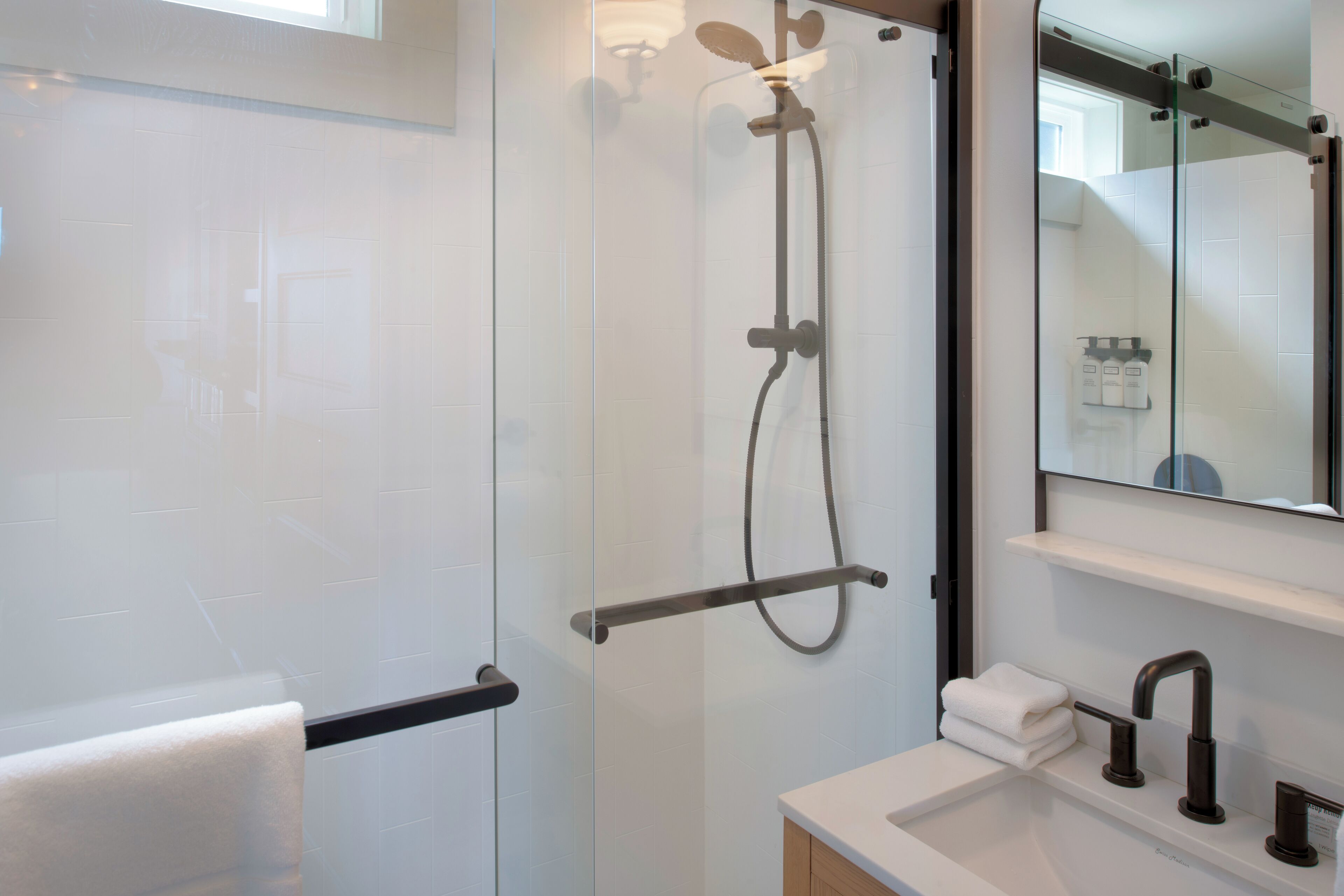 Une salle de bain moderne avec une douche à parois de verre, un lavabo blanc doté d’un robinet foncé et un miroir au-dessus du lavabo.