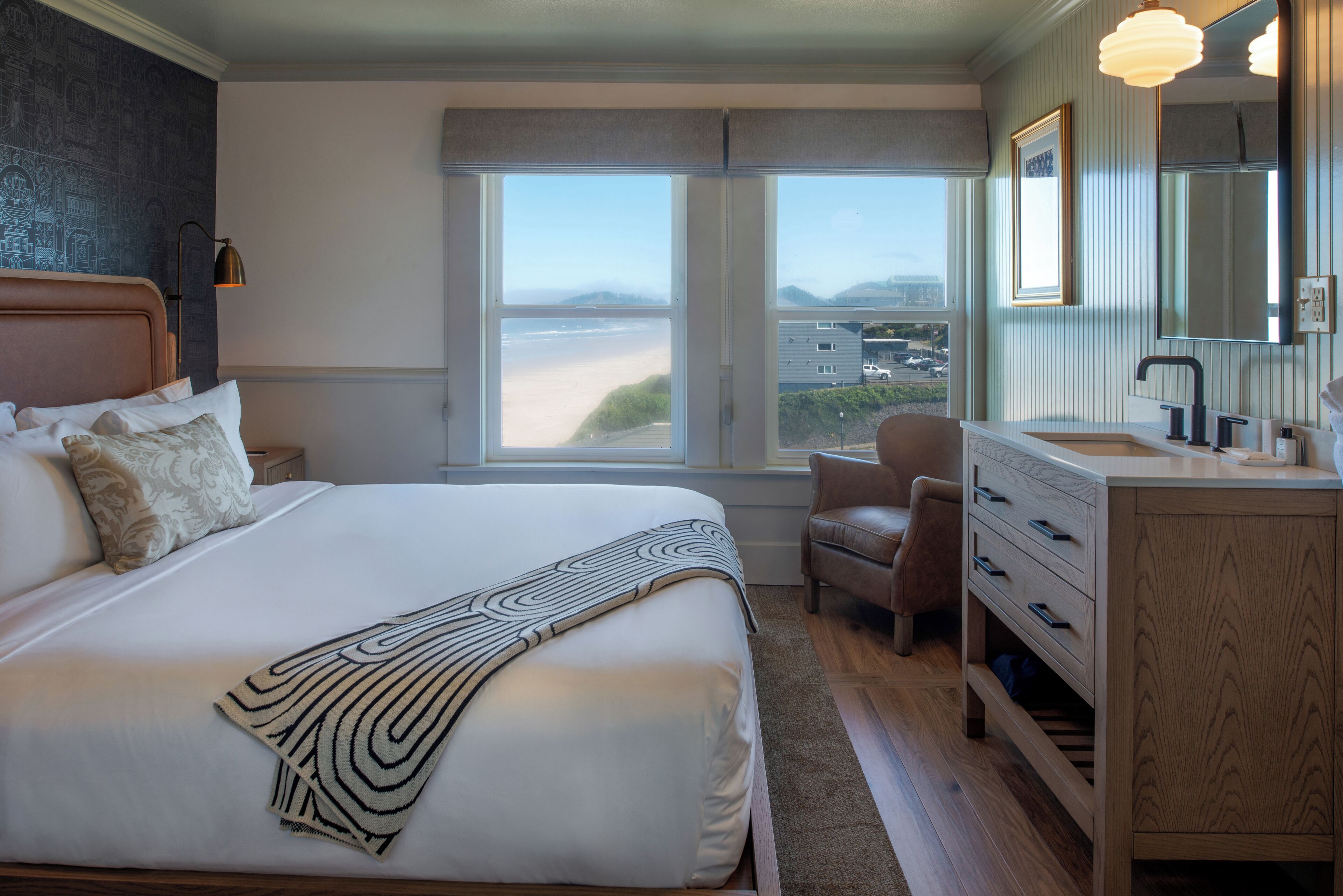 A bedroom with a large bed, a view of the beach, a chair, and a wooden dresser with drawers.