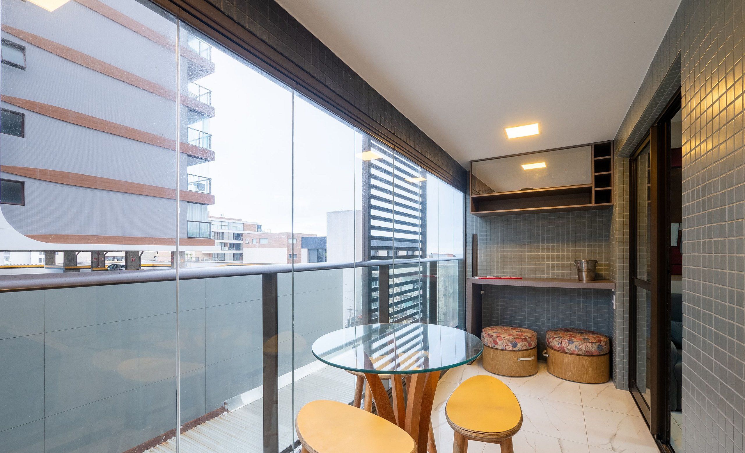 Un balcon avec une table en verre et deux chaises, un petit coin salon avec des tabourets et une vue sur les immeubles.