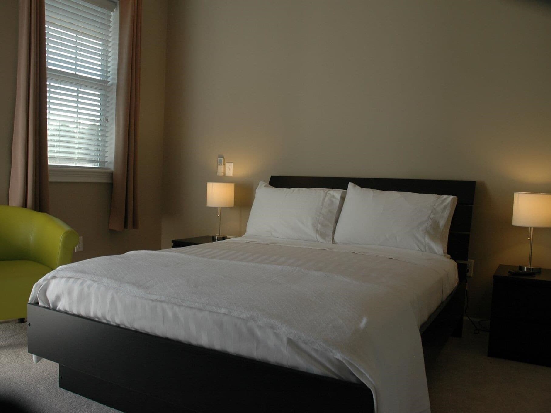 Habitación de hotel con una cama grande, dos lámparas de noche, una mesita de noche, un sillón verde y una ventana con persianas.