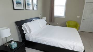 Una cama bien hecha en una habitación de hotel, con cabecera negra, mesita de noche con lámpara y una silla.