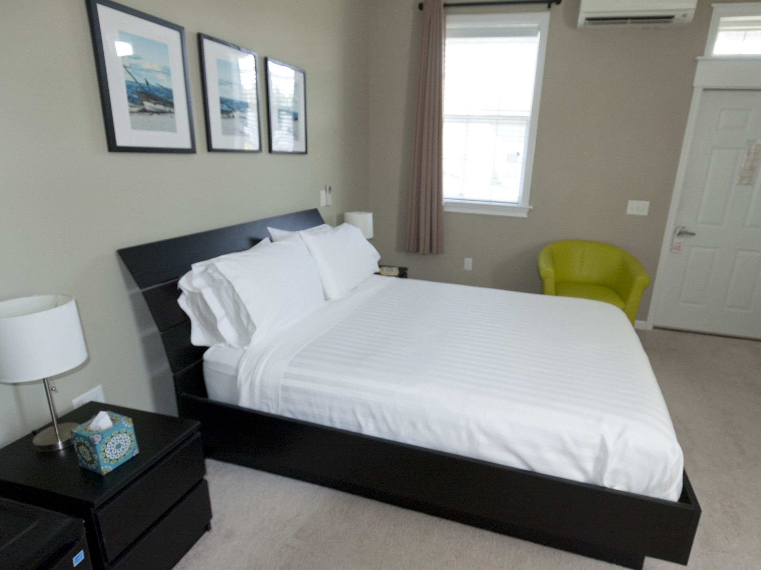 A neatly made bed in a hotel room with a black headboard, a bedside table with a lamp, and a chair.
