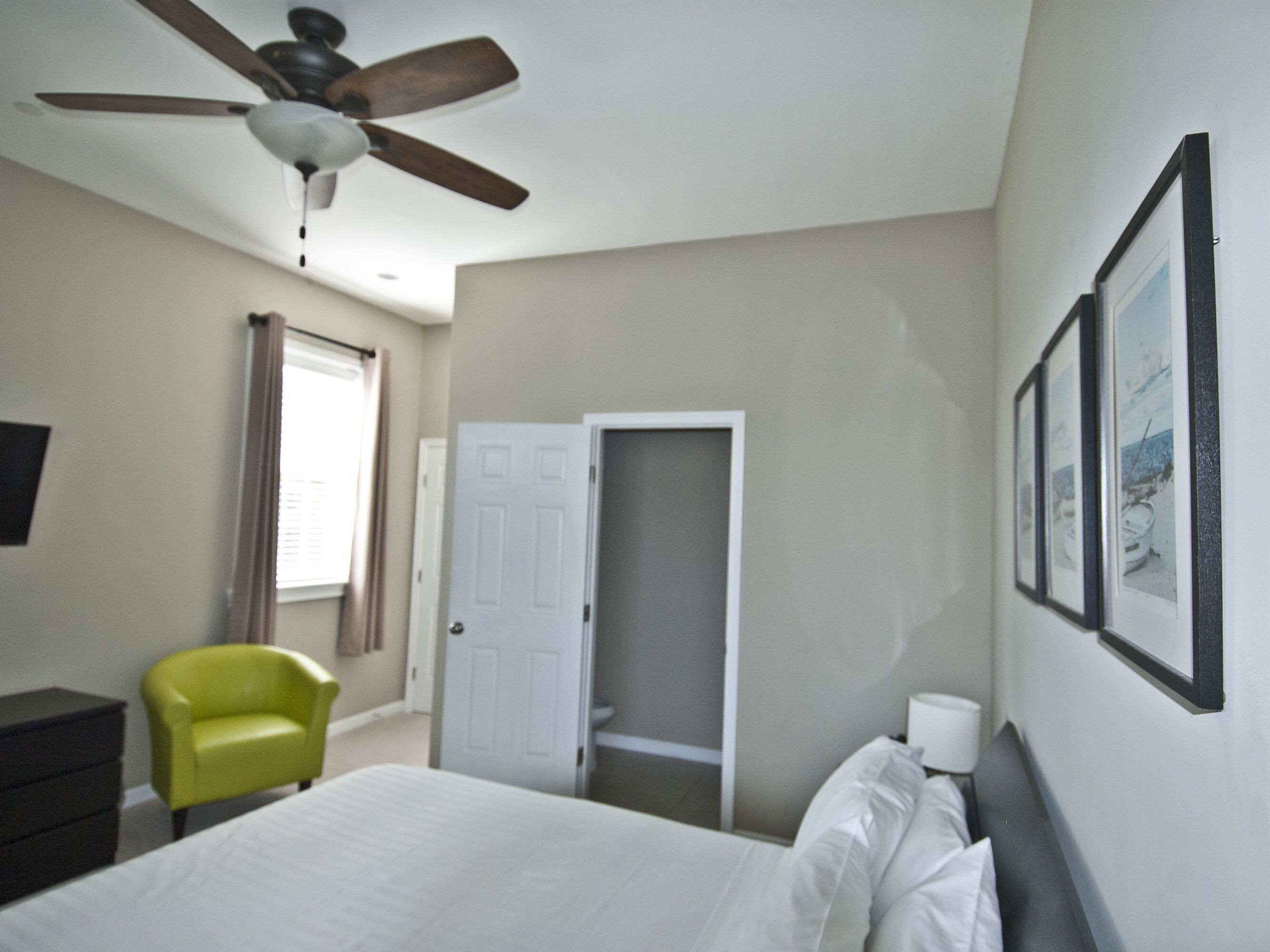 A bedroom with a bed, a ceiling fan, a chair, a door, and framed pictures on the wall.