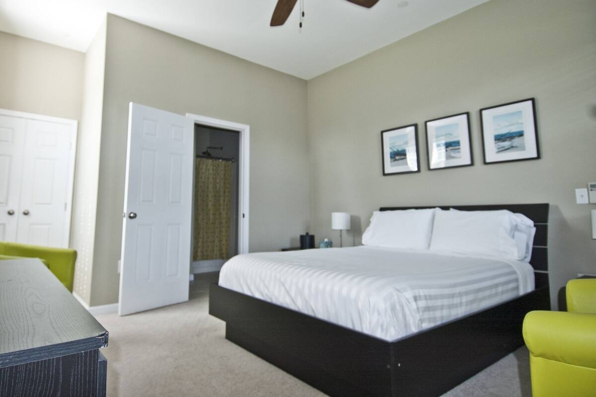 A bedroom with a bed, a ceiling fan, a door, a chair, and a wall with three framed pictures.