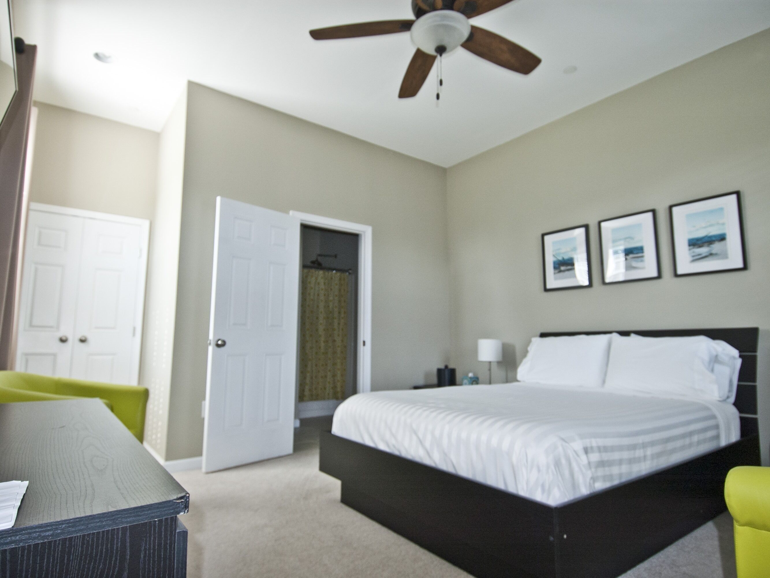 Quarto de hotel com cama, escrivaninha, cadeira, ventoinha no teto e porta.