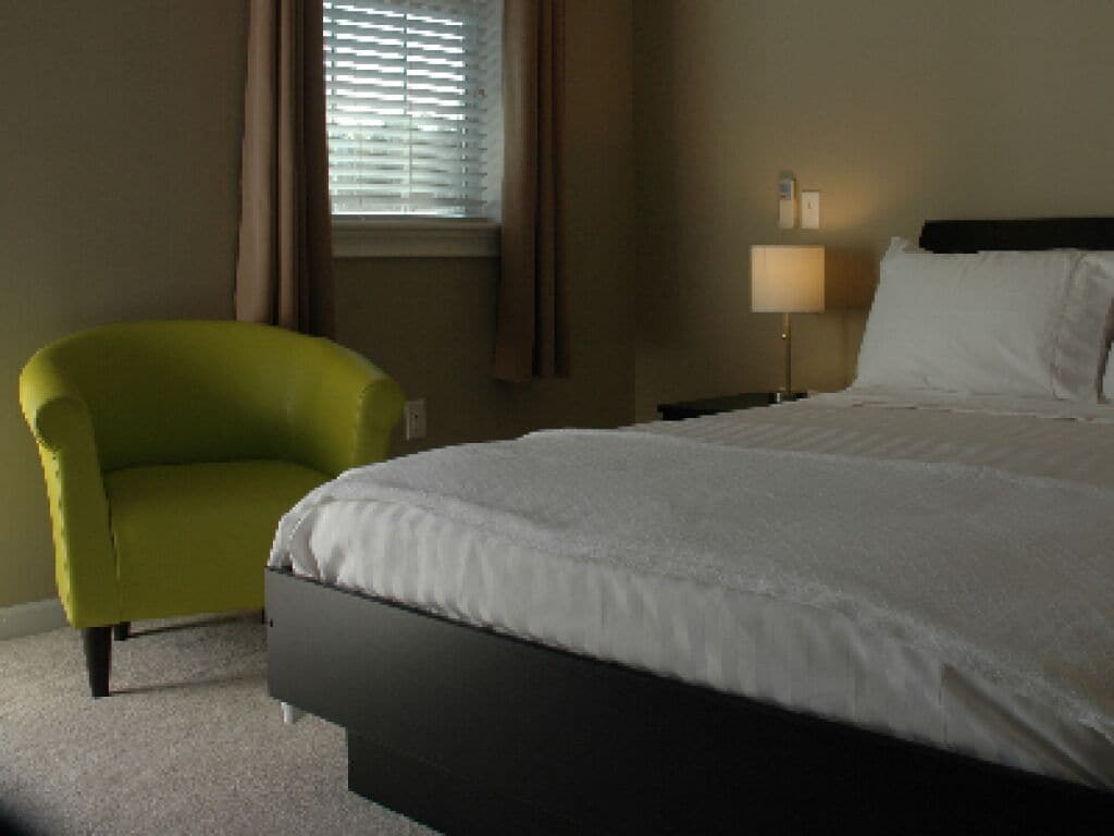 Quarto de hotel com cama, poltrona, luminária e janela com cortinas.