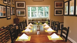 Un comedor con una larga mesa de madera dispuesta para seis comensales, rodeada de sillas. Hay cuadros en las paredes, una ventana con vistas a árboles y un sofá al fondo.