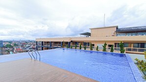 A rooftop swimming pool with a wooden deck, surrounded by a building with multiple balconies and a view of the cityscape.