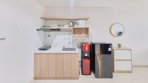 A compact kitchen with a marble countertop, wooden cabinets, a coffee maker, a kettle, a refrigerator, and a small plant. - Simply Look 1Br Apartment Patraland Amarta (Ngaglik)