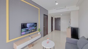 A modern living room with a flat-screen TV mounted on the wall, a small round table with two bottles, a grey sofa, and a wooden cabinet. - Well Furnished 1Br At Patraland Amarta Apartment (Ngaglik)