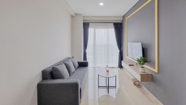A modern living room with a grey sofa, a small round table, a TV on a wall-mounted stand, and a window with sheer curtains. - Well Furnished 1Br At Patraland Amarta Apartment (Ngaglik)