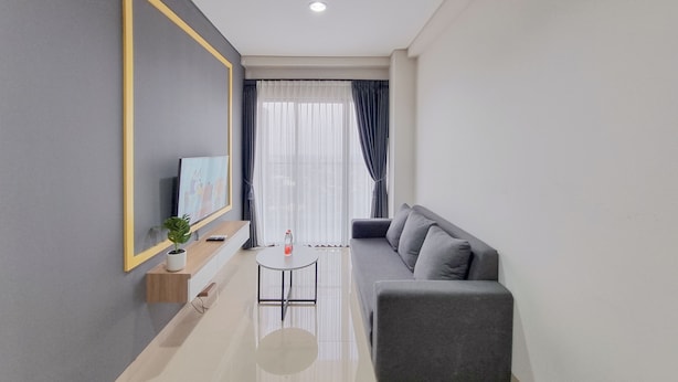 Ein modernes Wohnzimmer mit einer grauen Couch, einem kleinen runden Tisch, einem an der Wand montierten Fernseher und einem Fenster mit Vorhängen.