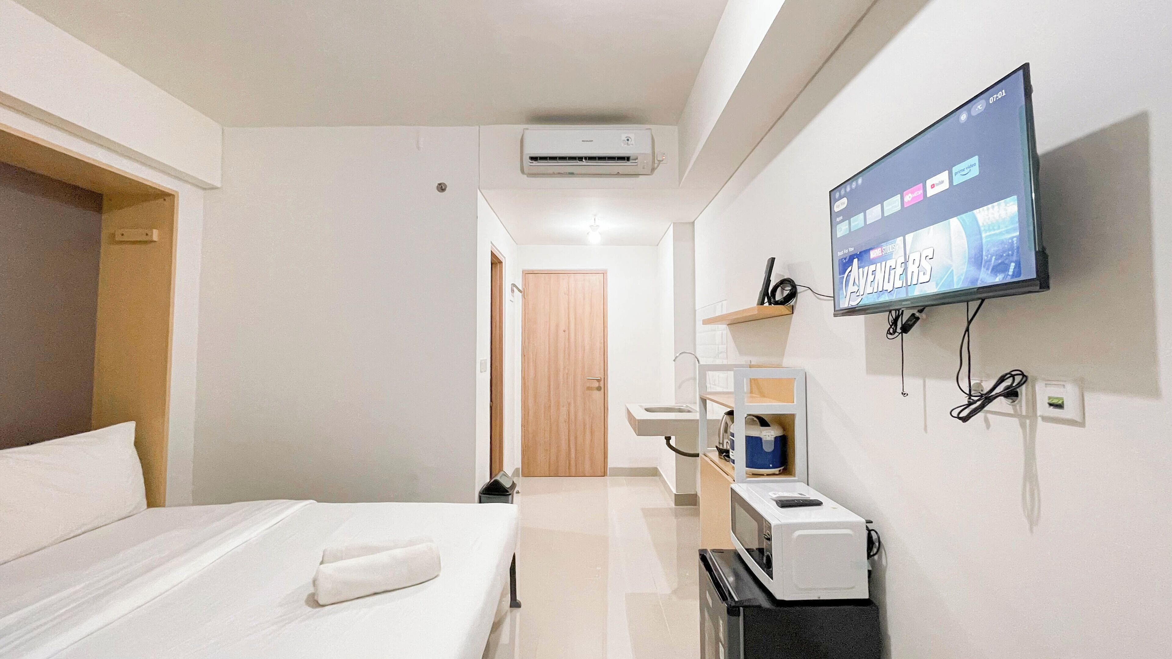 Habitación de hotel con una cama, un televisor mostrando 'Avengers', un microondas, un lavabo y una puerta que conduce a otra habitación.