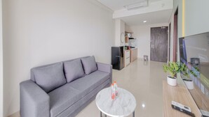 A modern living room with a grey sofa, a small round coffee table with two bottles, a remote control, and a TV on a wooden stand. - Spacious Apartment 1Br At Patraland Amarta (Ngaglik)